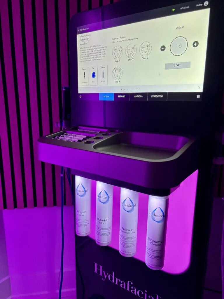 A HydraFacial device beside a stack of serum bottles, its touchscreen lit under purple light