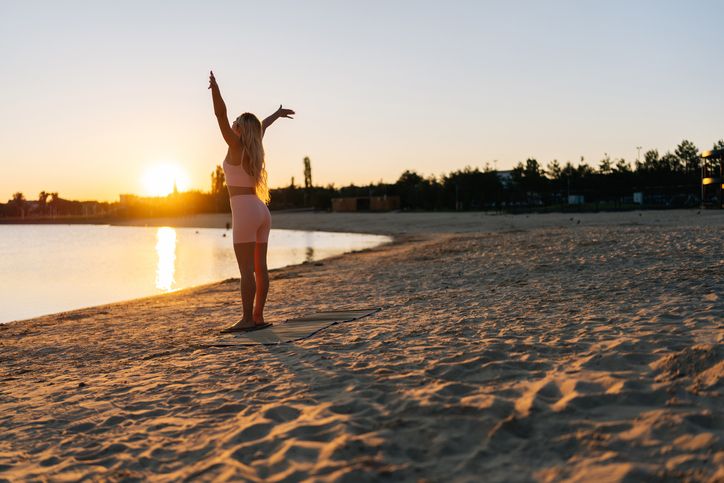 A woman stretching with both arms raised on a beach at sunrise