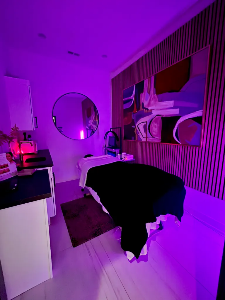 A treatment bed in a spa room with purple lighting, wall art, cabinetry, and a circular mirror on the wall.